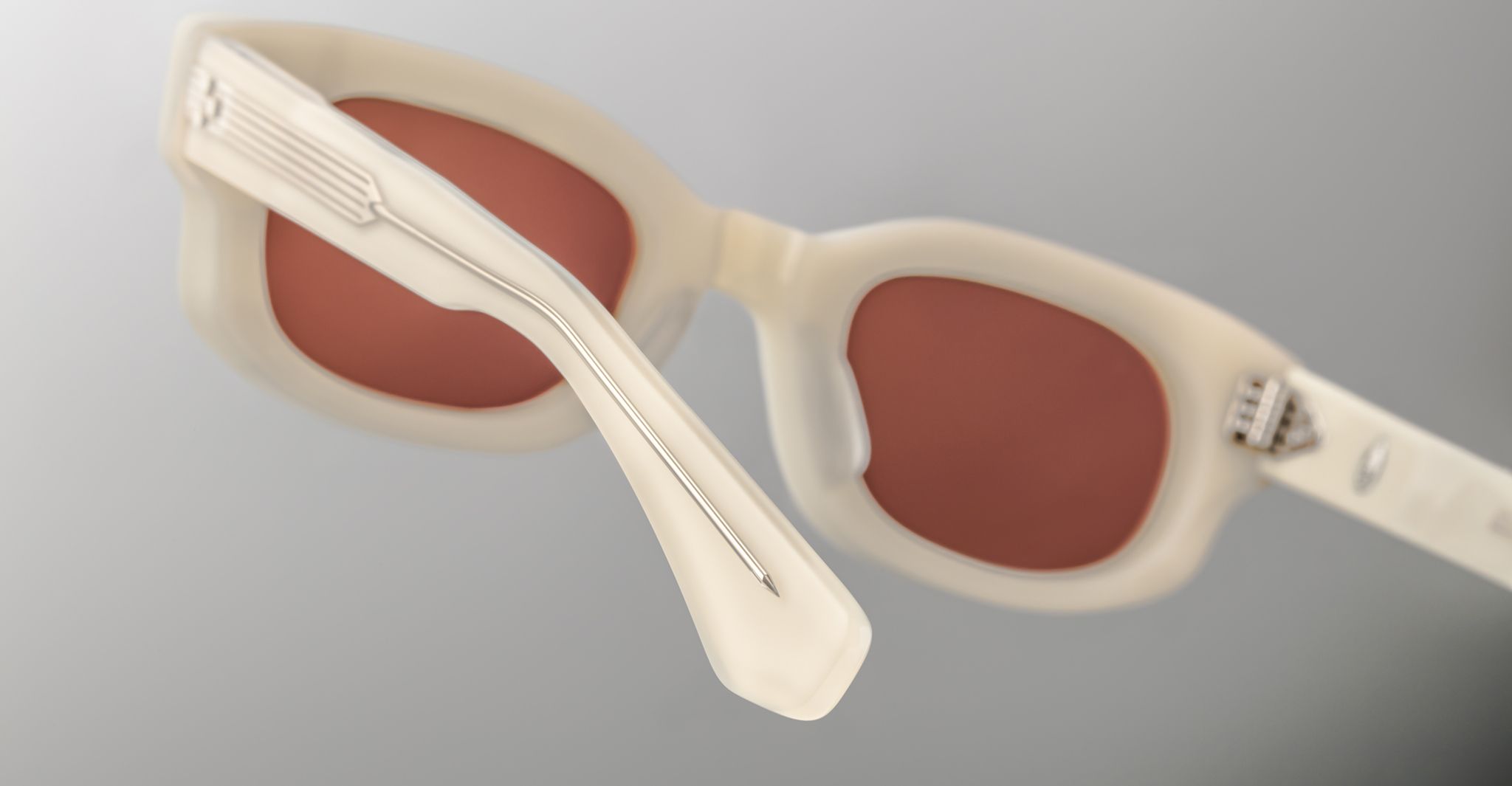 Close-up of Jacques Marie Mage Whiskeyclone sunglasses featuring thick, off-white frames and reddish-brown lenses, set against a gray gradient background. The image emphasizes the detailed temples and lens craftsmanship.