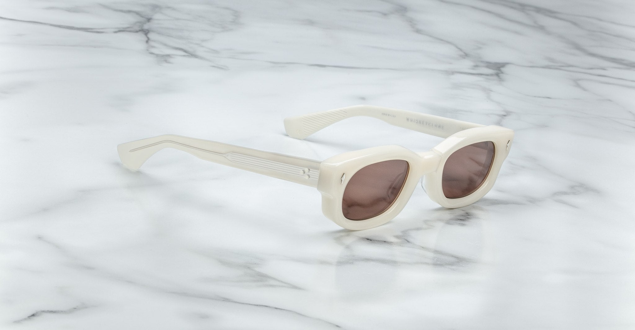Jacques Marie Mage Whiskeyclone sunglasses with cream-colored, round tinted lenses and thick frames rest on a white marble surface.