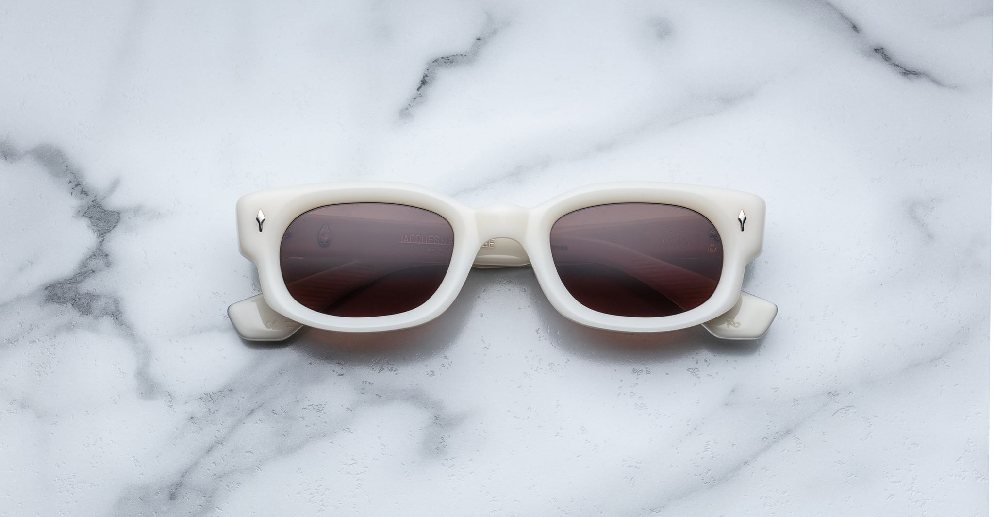 Jacques Marie Mage Whiskeyclone sunglasses with white frames and dark lenses are showcased on a white marble surface with grey veining.