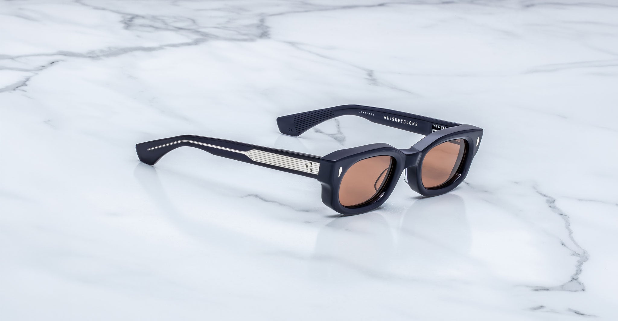 Jacques Marie Mage Whiskeyclone in Minuit sunglasses, deep navy blue acetate frame with warm rose-tinted lenses, signature silver-tone arrowhead rivets, and engraved silver-tone metal core temples with three-dot detailing, shown in a three-quarter angled view on a white marble surface