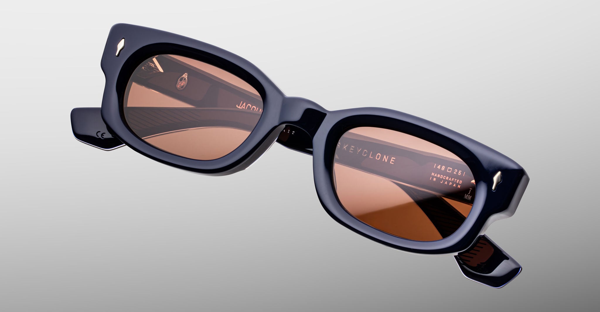 Jacques Marie Mage Whiskeyclone in Minuit sunglasses, deep navy blue acetate frame with warm rose-tinted lenses, sculpted rectangular silhouette, and signature silver-tone arrowhead rivets, shown in an angled front close-up highlighting the bold frame design and visible inner temple detailing