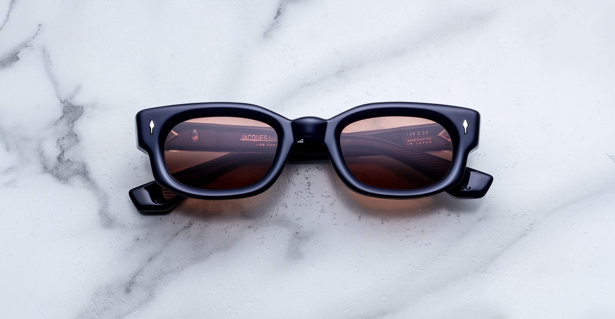 Jacques Marie Mage Whiskeyclone in Minuit sunglasses, deep navy blue acetate frame with warm rose-tinted lenses, bold rectangular silhouette, sculpted bridge, and signature silver-tone arrowhead rivets, shown in a straight-on front view against a white marble background