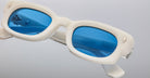 Close-up of white-framed Jacques Marie Mage Whiskeyclone sunglasses with bright blue tinted lenses, reflecting light and featuring small metallic details. The brand name is visible on the inside of the left lens.