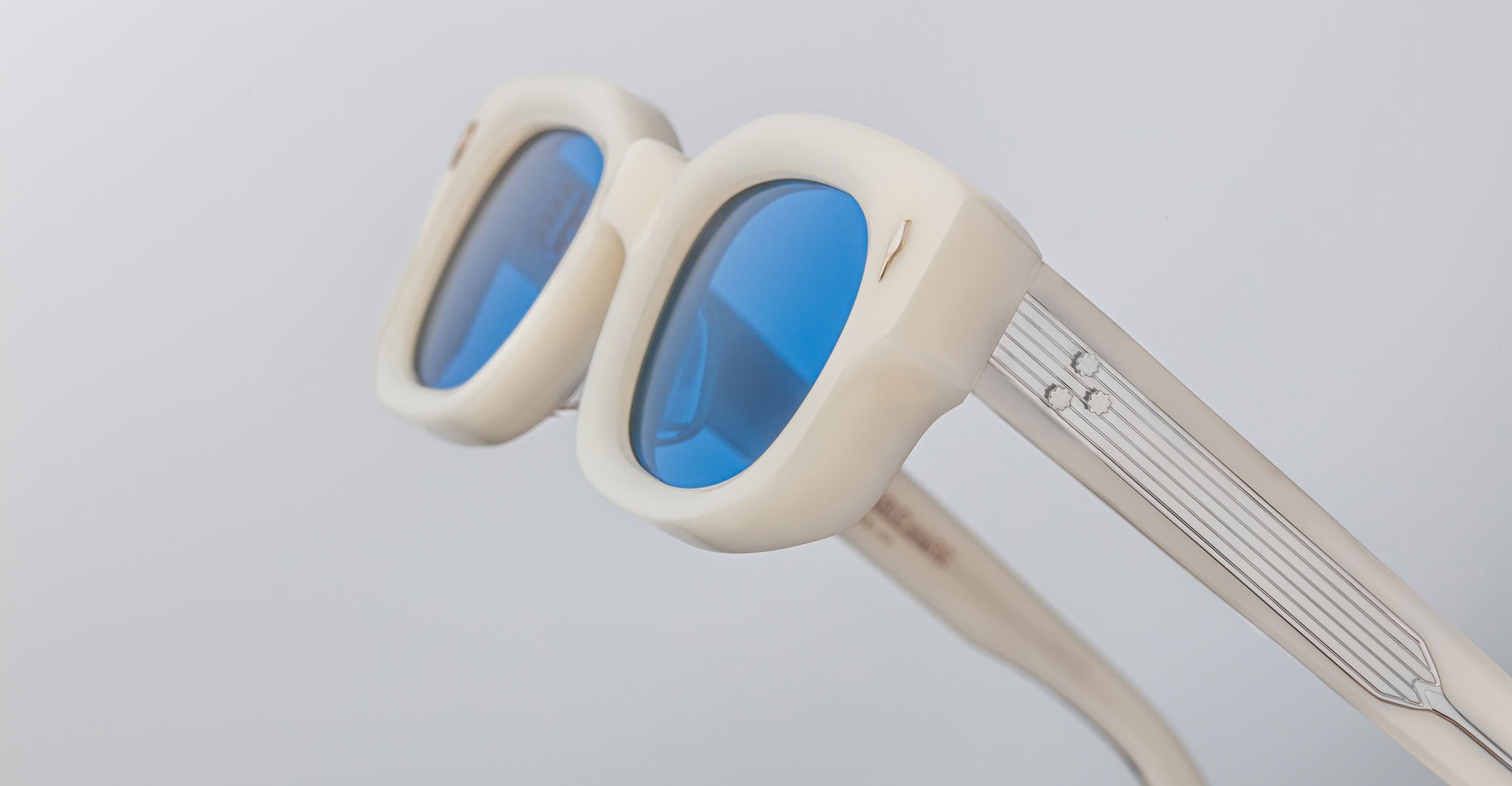 Close-up of Jacques Marie Mage Whiskeyclone sunglasses with thick cream frames, blue-tinted lenses, silver striped arms, and a small decorative detail, set against a light gray background.
