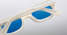 Close-up of Jacques Marie Mage Whiskeyclone sunglasses with white frames and blue lenses, viewed at an angle from above. Partially folded arms display metal accents against a plain light gray background.