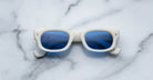 Jacques Marie Mage Whiskeyclone sunglasses, featuring a rectangular white frame and blue lenses, rest facing upward on a white marble surface, with the lenses reflecting hints of the surrounding environment.