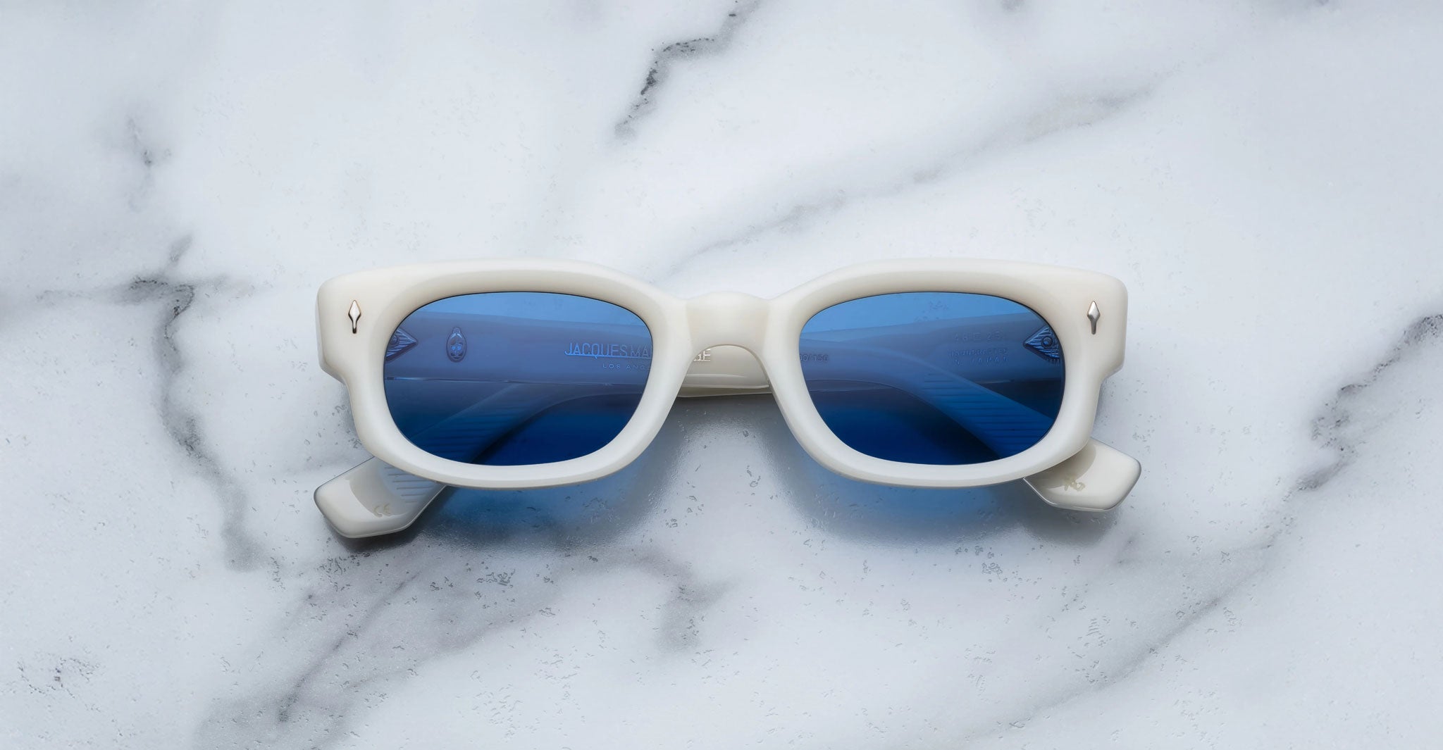 Jacques Marie Mage Whiskeyclone sunglasses, featuring a rectangular white frame and blue lenses, rest facing upward on a white marble surface, with the lenses reflecting hints of the surrounding environment.