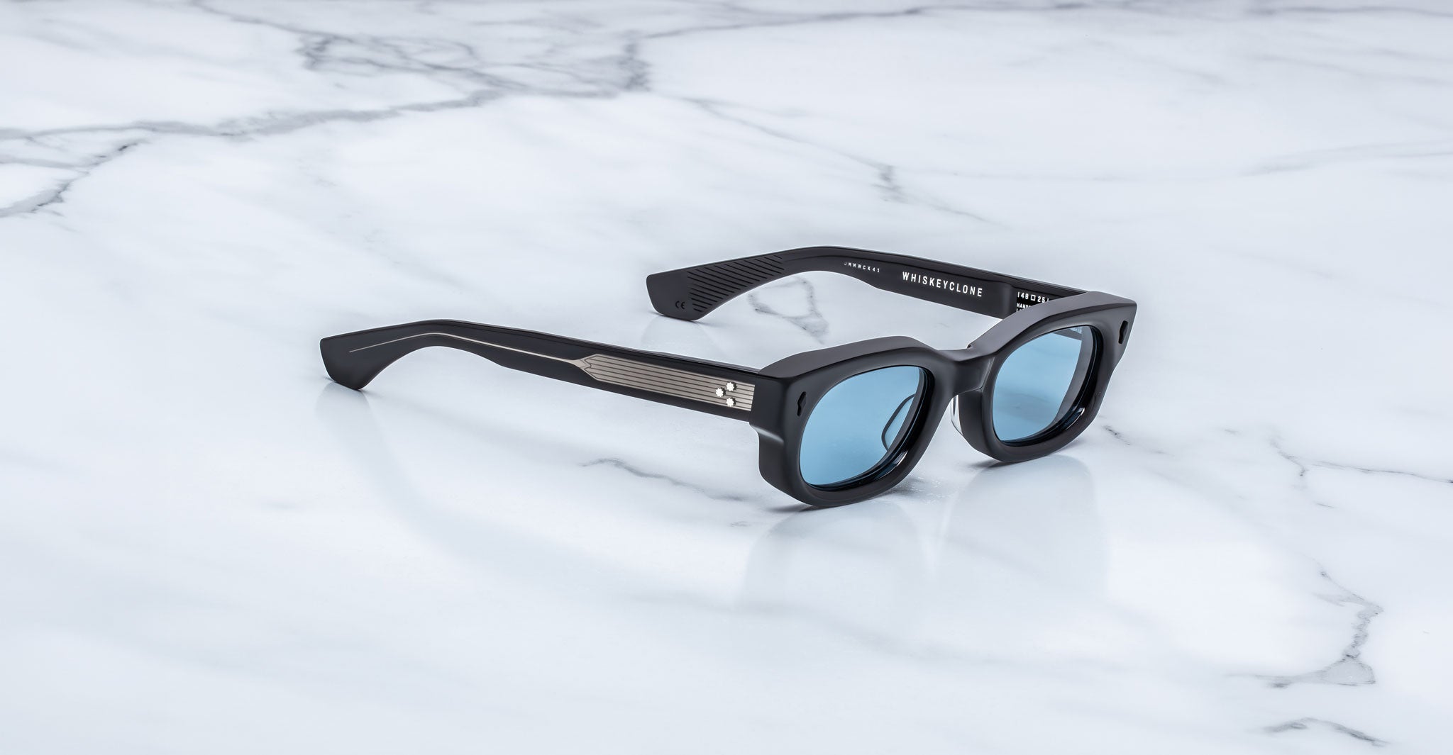 Jacques Marie Mage Whiskeyclone in Mystique sunglasses, dark charcoal acetate frame with light blue-tinted lenses, signature black arrowhead rivets, and engraved silver-tone metal core temples with three-dot detailing, shown in a three-quarter angled view on a white marble surface