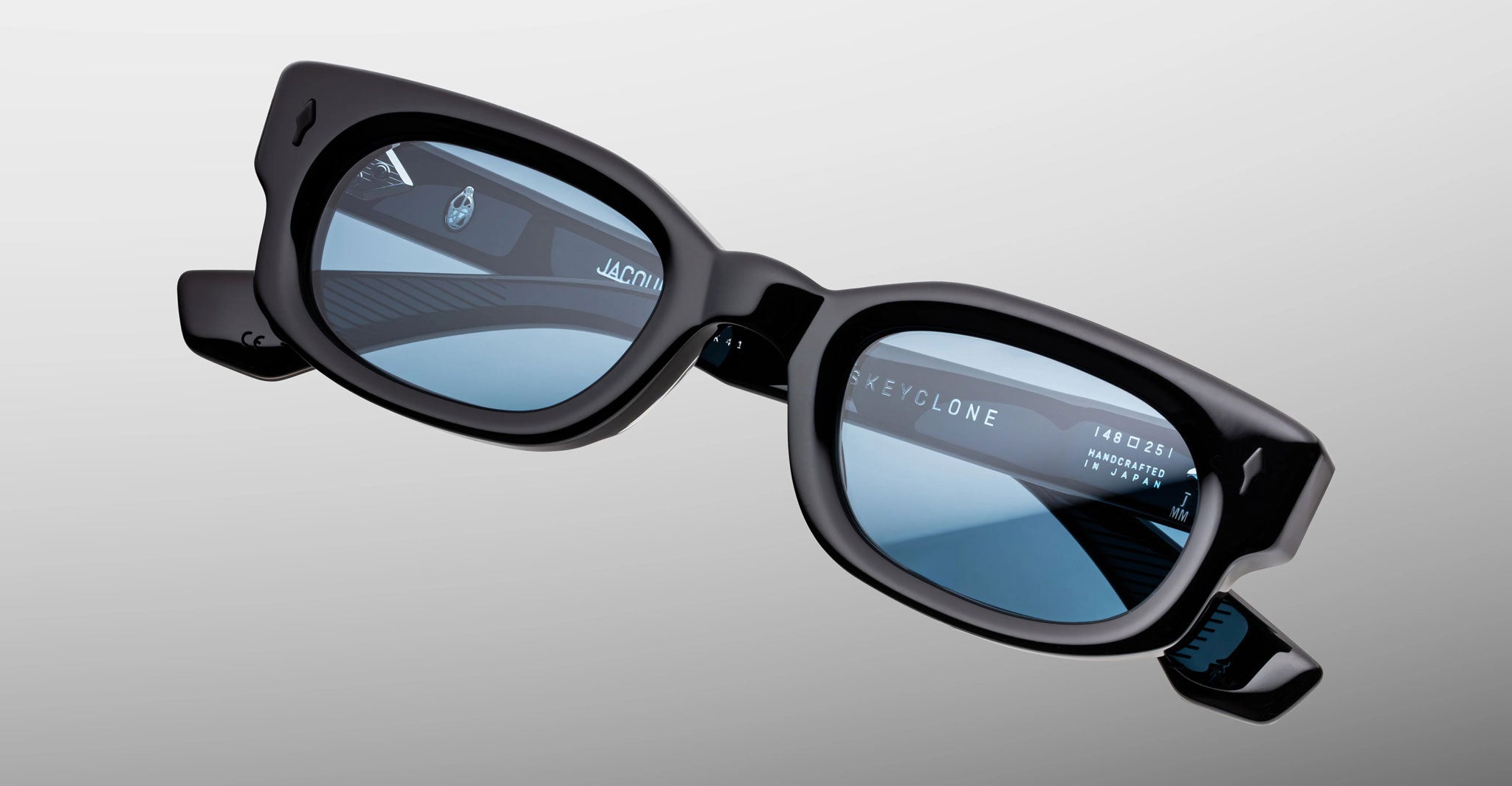 Jacques Marie Mage Whiskeyclone in Mystique sunglasses, dark charcoal acetate frame with light blue-tinted lenses, bold rectangular silhouette, sculpted bridge, and signature black arrowhead rivets, shown in an angled front close-up highlighting the sculptural frame design and visible inner temple detailing