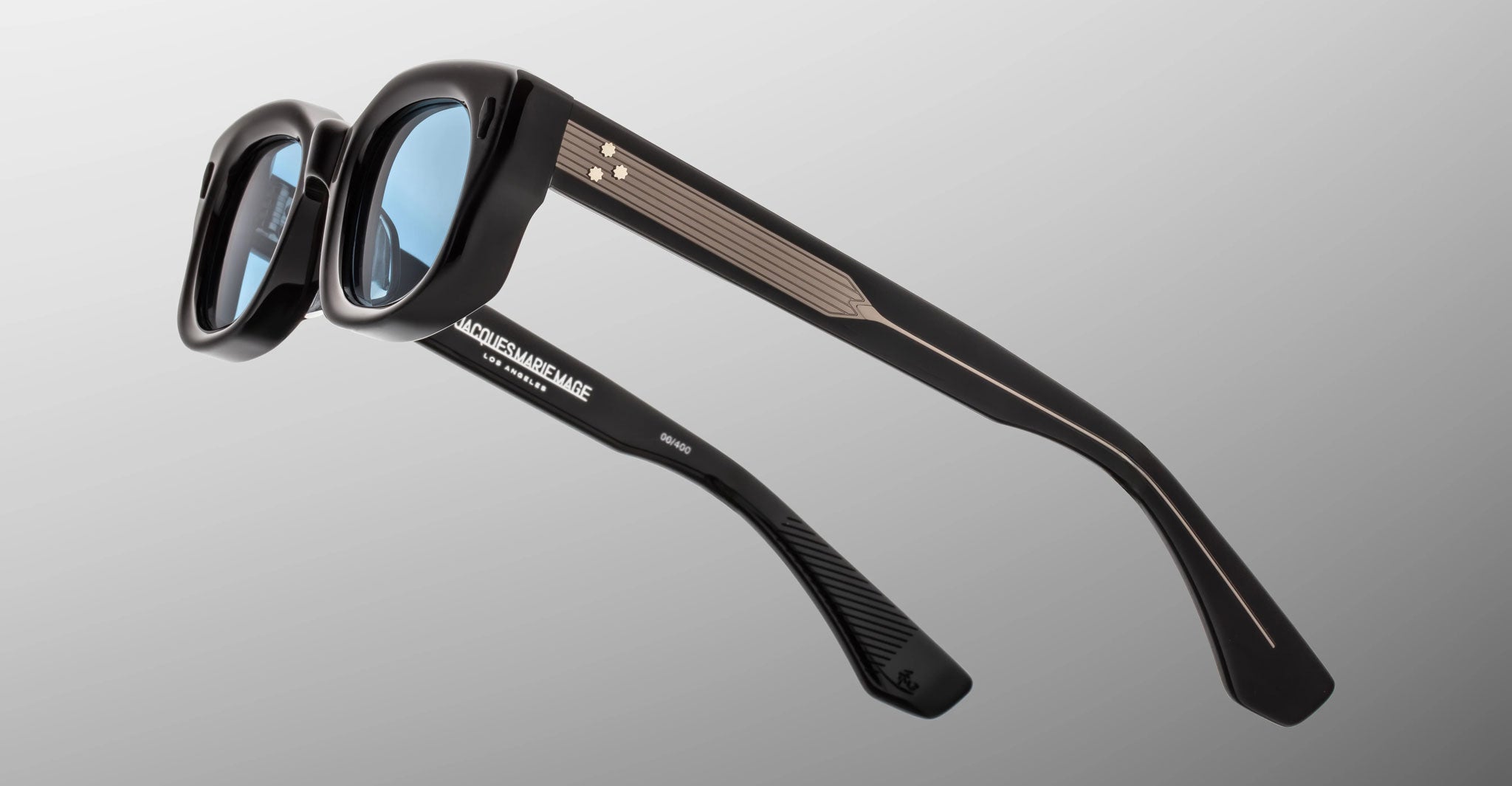 Jacques Marie Mage Whiskeyclone in Mystique sunglasses, dark charcoal acetate frame with light blue-tinted lenses, signature silver-tone three-dot rivets, and engraved silver-tone metal core temples, shown from a rear side angle highlighting the sculpted frame and Jacques Marie Mage branding on the inner temple