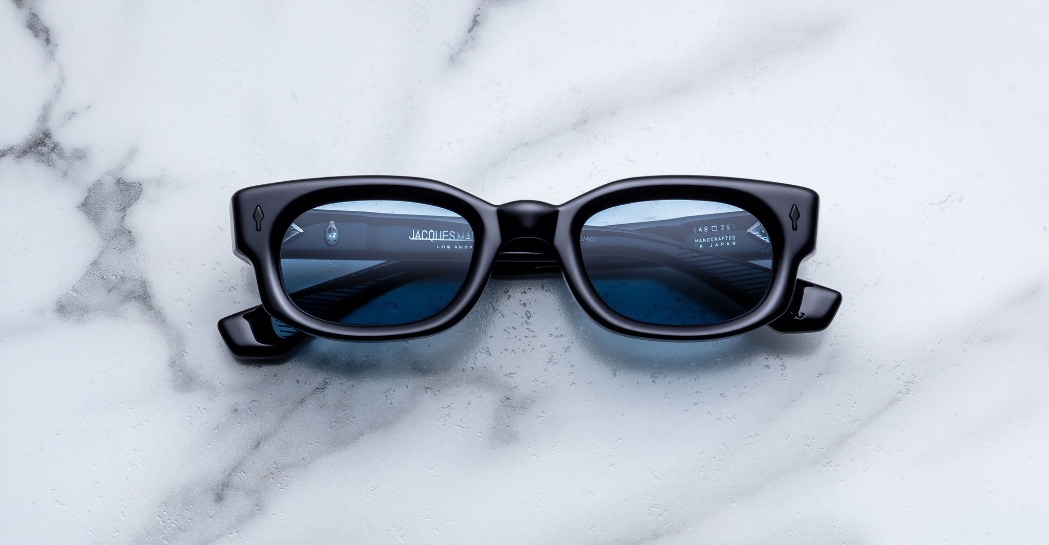 Jacques Marie Mage Whiskeyclone in Mystique sunglasses, dark charcoal acetate frame with blue-tinted lenses, bold rectangular silhouette, sculpted bridge, and signature black arrowhead rivets, shown in a straight-on front view against a white marble background
