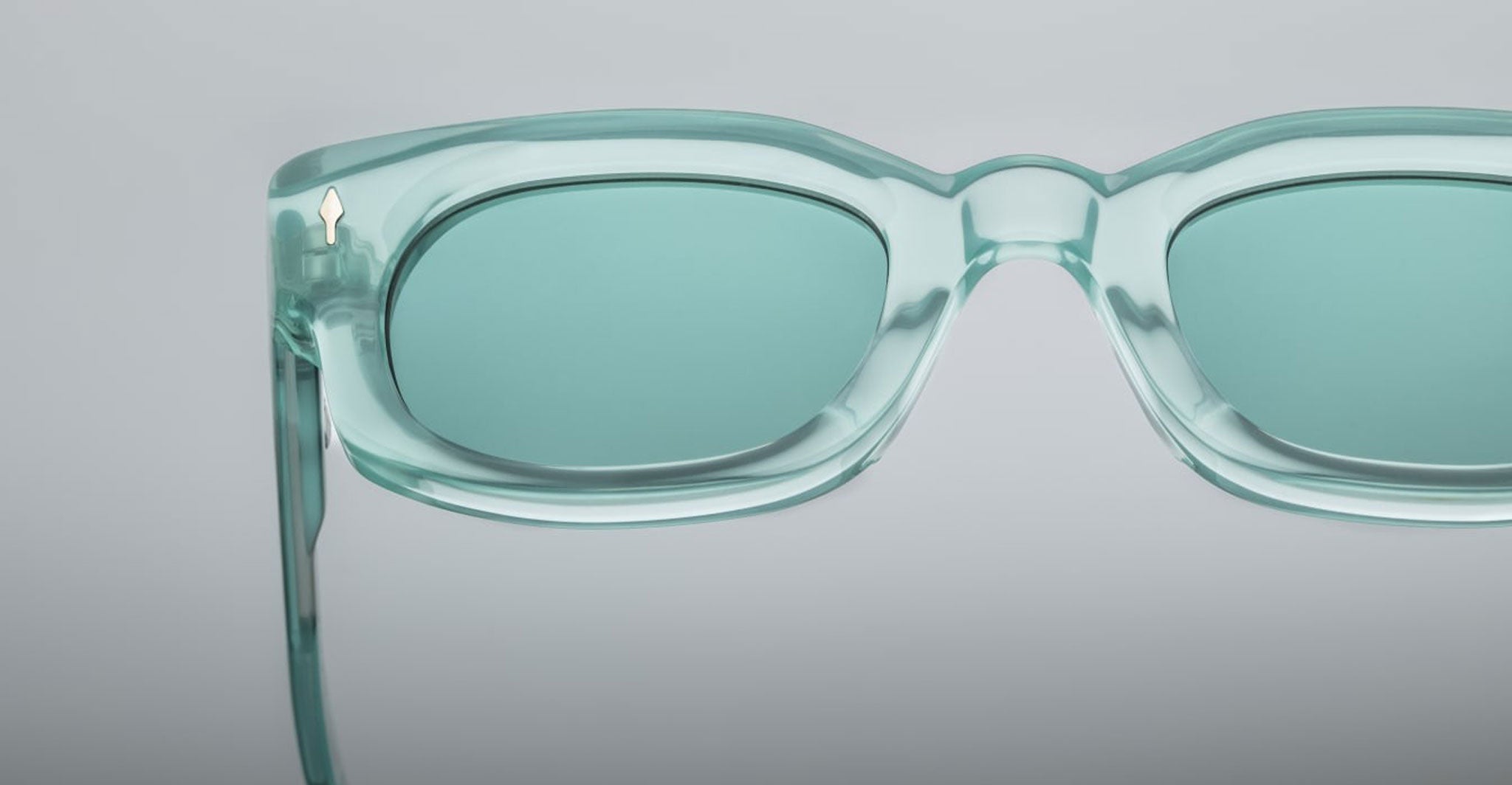 Close-up of Jacques Marie Mage Whiskeyclone sunglasses in translucent teal, featuring rectangular frames and matching tinted lenses. Set on a plain light gray background with only the top half and right side of the frame visible.