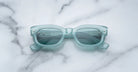 A pair of Jacques Marie Mage Whiskeyclone rectangular, light blue sunglasses with dark lenses rests on a white marble surface featuring grey veining.