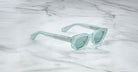The Jacques Marie Mage Whiskeyclone sunglasses, in light blue translucent with thick frames and matching tinted lenses, are displayed on a white marble surface with gray veining.