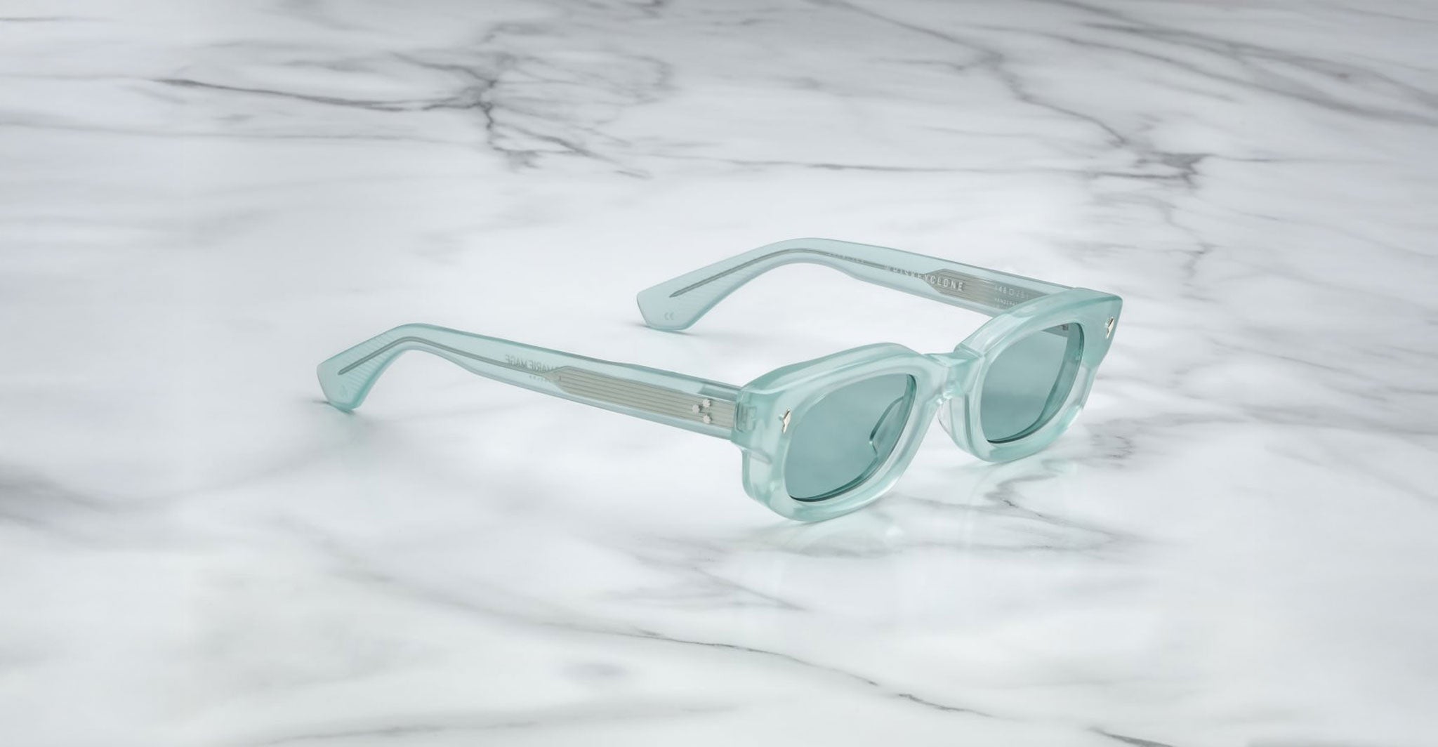 The Jacques Marie Mage Whiskeyclone sunglasses, in light blue translucent with thick frames and matching tinted lenses, are displayed on a white marble surface with gray veining.