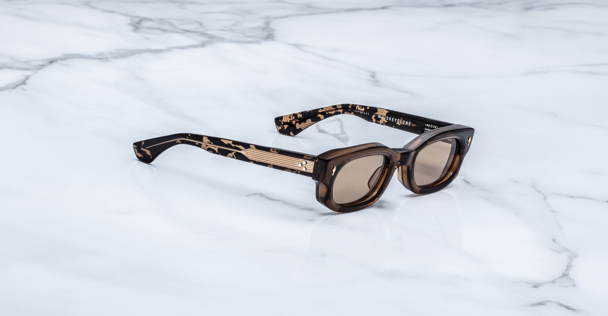 Jacques Marie Mage Whiskeyclone in Tawny sunglasses, translucent brown acetate frame with warm taupe-tinted lenses, signature gold-tone arrowhead rivets, and patterned tortoiseshell temples featuring engraved gold-tone metal cores and three-dot detailing, shown in a three-quarter angled view on a white marble surface