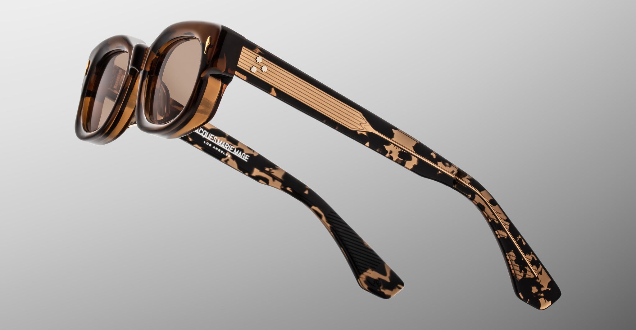Jacques Marie Mage Whiskeyclone in Tawny sunglasses, translucent brown tortoiseshell acetate frame with warm taupe-tinted lenses, signature gold-tone arrowhead rivets, and engraved gold-tone metal core temples with three-dot detailing, shown from a rear side angle highlighting the patterned acetate and Jacques Marie Mage branding on the inner temple