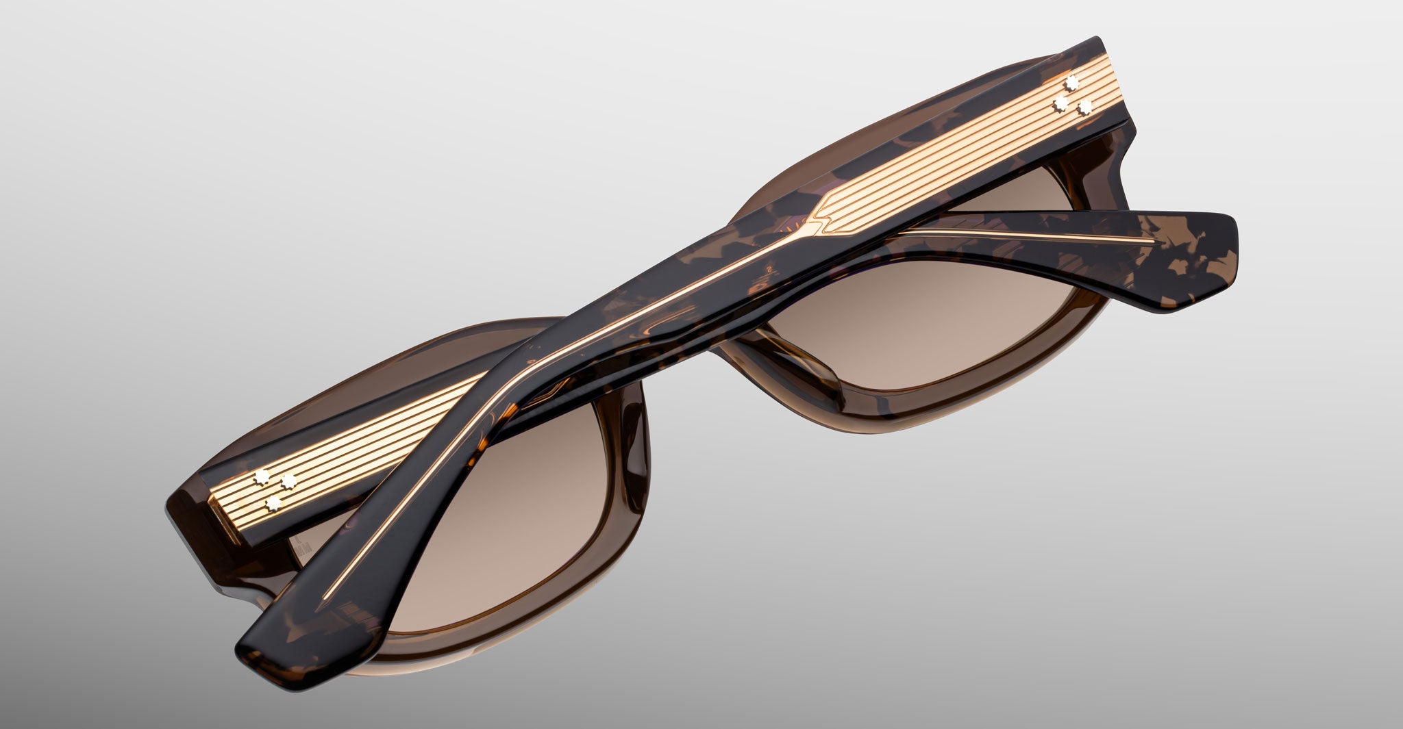 Jacques Marie Mage Whiskeyclone in Tawny sunglasses, translucent brown tortoiseshell acetate frame with warm taupe-tinted lenses and intricately engraved gold-tone metal core temples featuring three-dot rivet detailing, shown folded in a close-up view highlighting the layered acetate pattern and decorative hardware
