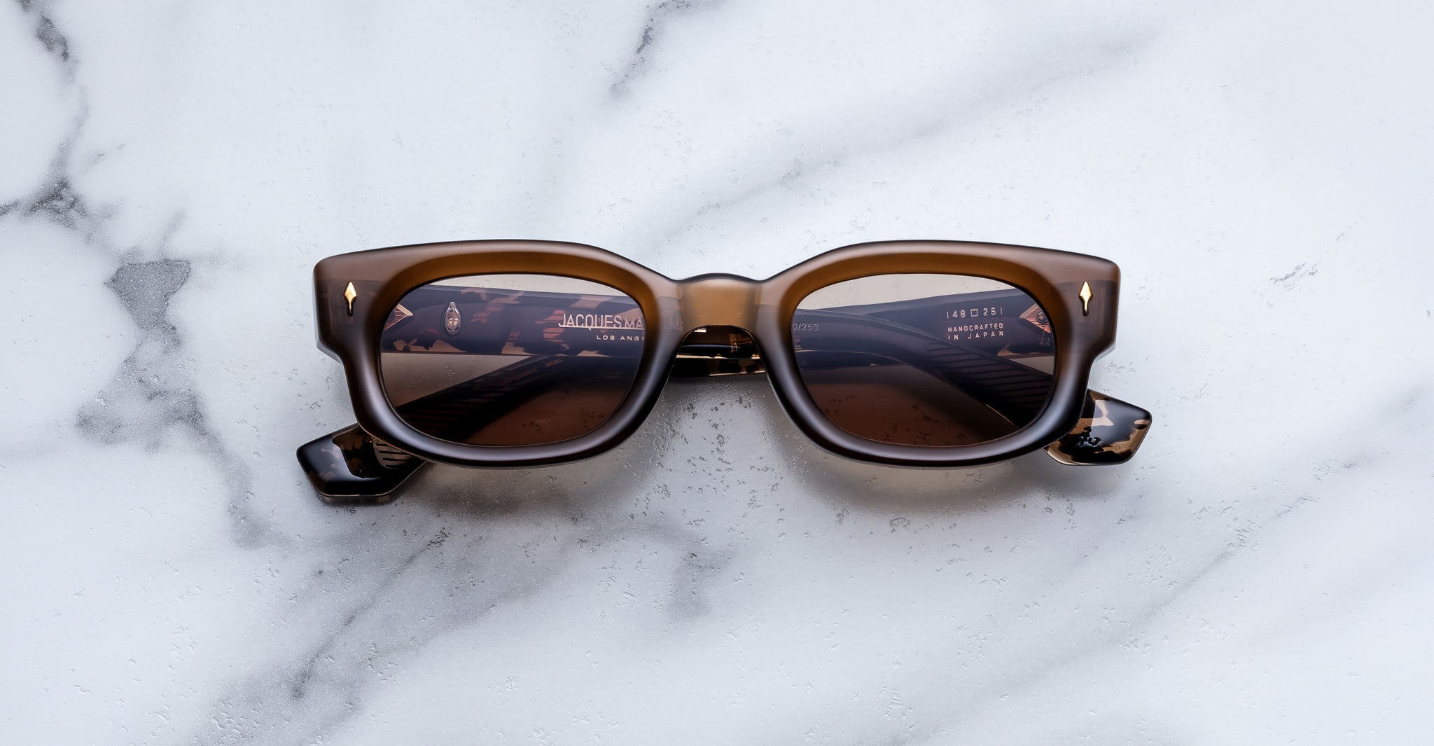 Jacques Marie Mage Whiskeyclone in Tawny sunglasses, translucent warm brown acetate frame with subtle tortoiseshell detailing, brown-tinted lenses, bold rectangular silhouette, sculpted bridge, and signature gold-tone arrowhead rivets, shown in a straight-on front view against a white marble background