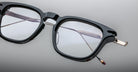 Close-up of Jacques Marie Mage William black rectangular eyeglasses with metal hinges and clear lenses, resting on a smooth light gray surface. The frame and temples are partially visible.