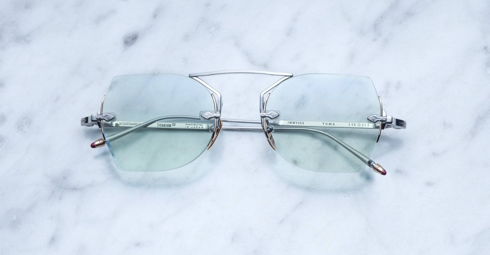 A pair of Jacques Marie Mage Yuma rimless sunglasses in Antique, featuring a metallic bridge frame and lightly tinted lenses, rests elegantly on a white marble surface with its sleek arms visible.