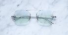 A pair of Jacques Marie Mage Yuma rimless sunglasses in Antique, featuring a metallic bridge frame and lightly tinted lenses, rests elegantly on a white marble surface with its sleek arms visible.