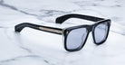 Jacques Marie Mage Yves black rectangular sunglasses with gold accents and dark polarized lenses, top-down view.