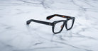 Jacques Marie Mage Yves in Noir 15 optical, rectangular black acetate frame with subtle tortoise interior, clear lenses, and signature arrowhead rivet detail, shown in angled side view on marble surface