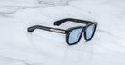 Jacques Marie Mage Yves in Obscure Black sunglasses, rectangular matte black acetate frame with blue-tinted lenses, signature arrowhead rivets, and engraved metal core temples, angled side view