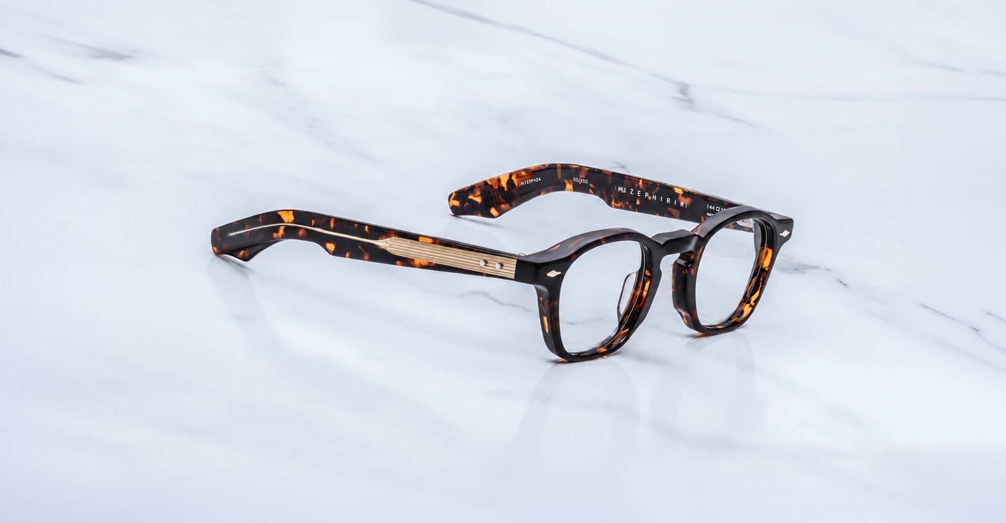 Jacques Marie Mage Zephirin optical frame in Aegen, three-quarter view with dark tortoiseshell acetate and gold-tone accents on a white marble surface.