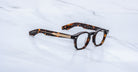Jacques Marie Mage Zephirin optical frame in Aegen, three-quarter view with dark tortoiseshell acetate and gold-tone accents on a white marble surface.
