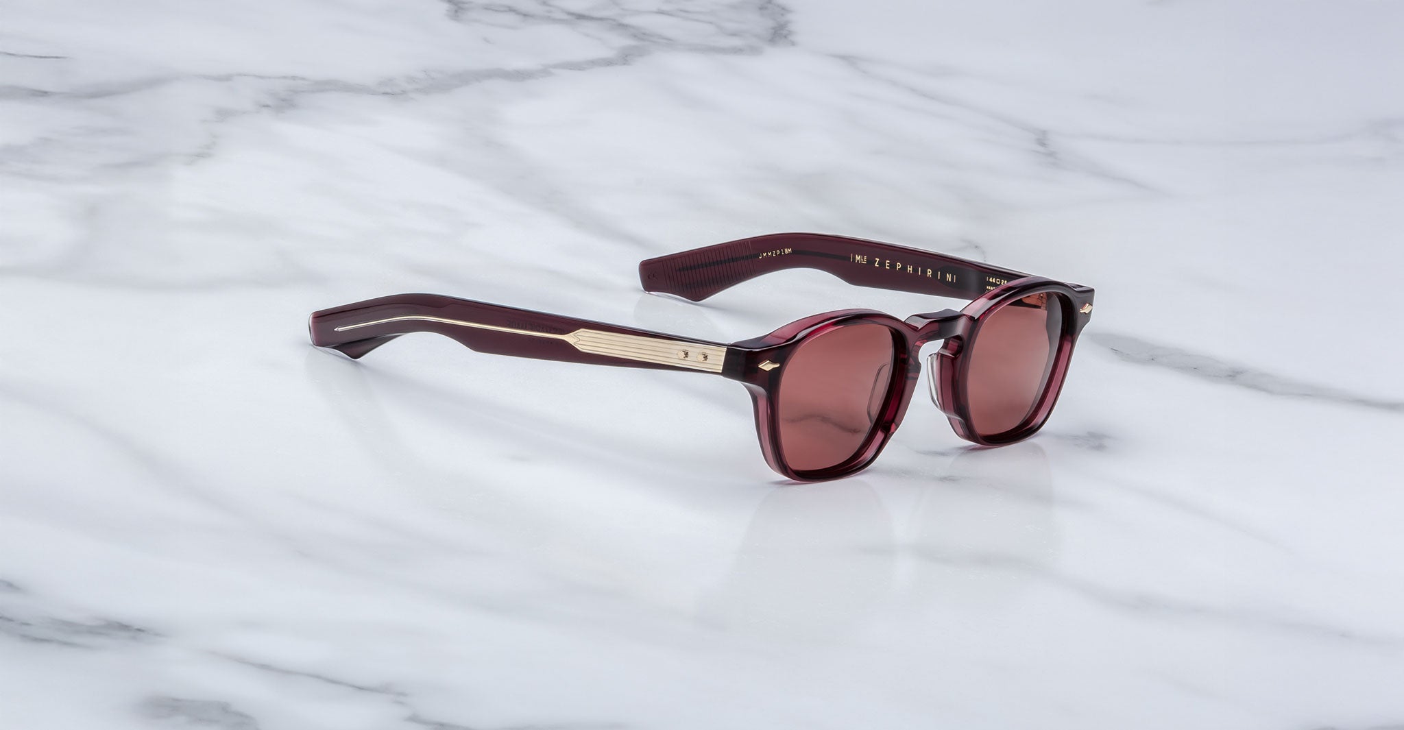 The Jacques Marie Mage Zephirin sunglasses feature maroon acetate frames, gold accents, and round tinted lenses. This handcrafted, limited-edition eyewear by Jacques Marie Mage rests elegantly on a white marble surface.