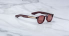 The Jacques Marie Mage Zephirin sunglasses feature maroon acetate frames, gold accents, and round tinted lenses. This handcrafted, limited-edition eyewear by Jacques Marie Mage rests elegantly on a white marble surface.