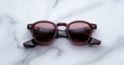 A pair of Jacques Marie Mage Zephirin sunglasses with round, burgundy frames and red-tinted lenses rests on a white marble surface—showcasing the brand’s exquisite limited edition eyewear.