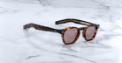 Limited edition Jacques Marie Mage Zephirin sunglasses, crafted from handcrafted acetate with a tortoiseshell pattern and round brown lenses, are displayed on a white marble surface.
