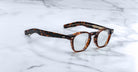 Jacques Marie Mage Zephirin spectacles by Jacques Marie Mage feature handcrafted acetate, round lenses, and a tortoiseshell finish, displayed on white marble to highlight the refined appeal of this limited edition eyewear.