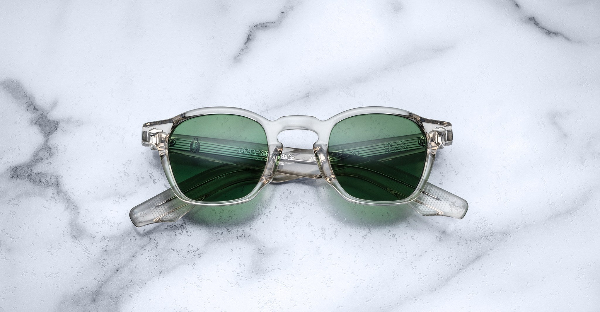 Jacques Marie Mage Zephirin sunglasses by Jacques Marie Mage, featuring clear frames and green lenses inspired by handcrafted acetate spectacles, are displayed folded on a white marble surface.