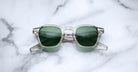 Jacques Marie Mage Zephirin sunglasses by Jacques Marie Mage, featuring clear frames and green lenses inspired by handcrafted acetate spectacles, are displayed folded on a white marble surface.
