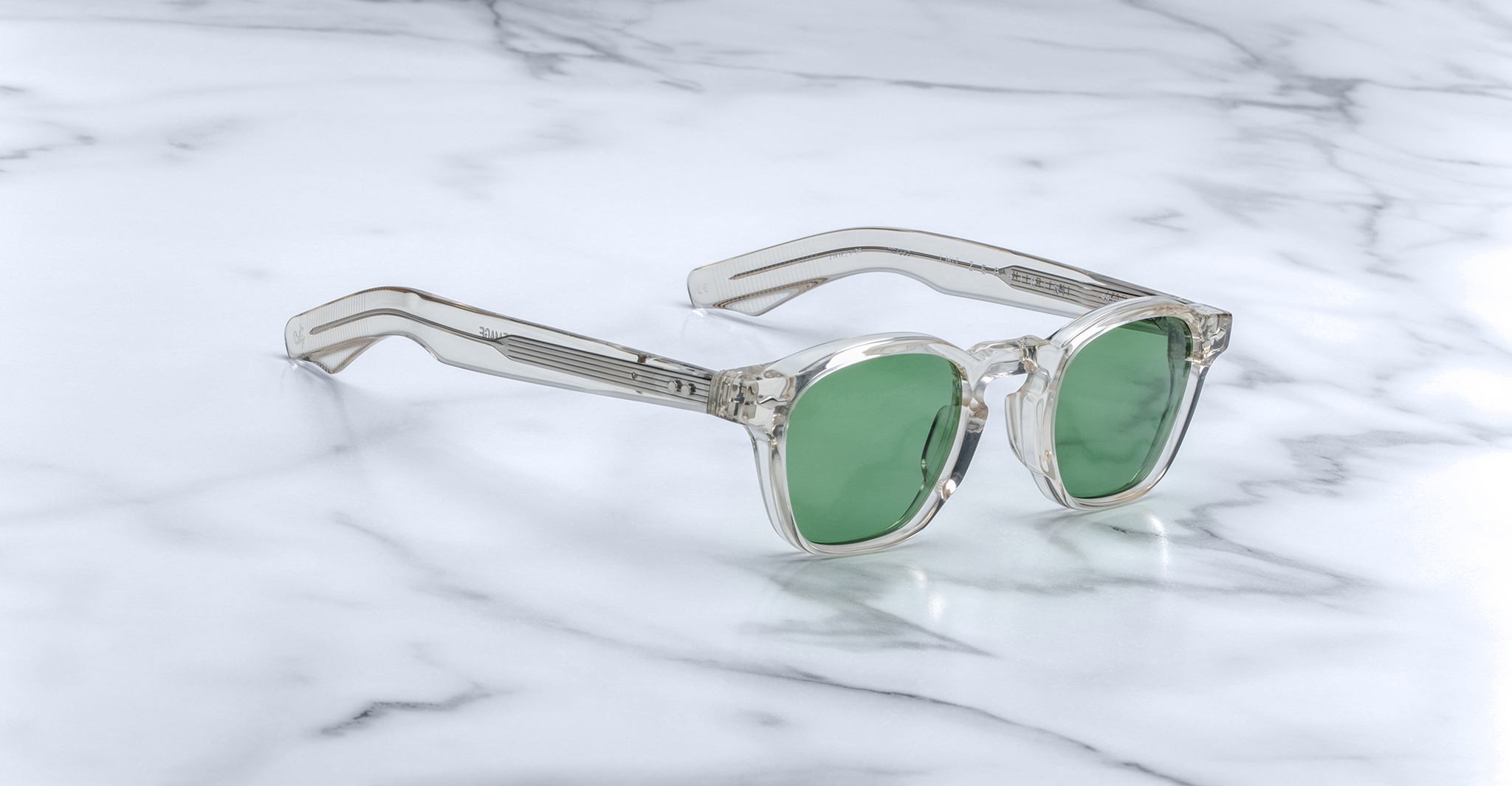 Jacques Marie Mage Zephirin sunglasses with clear acetate frames and green lenses by Jacques Marie Mage are displayed on a white marble surface.