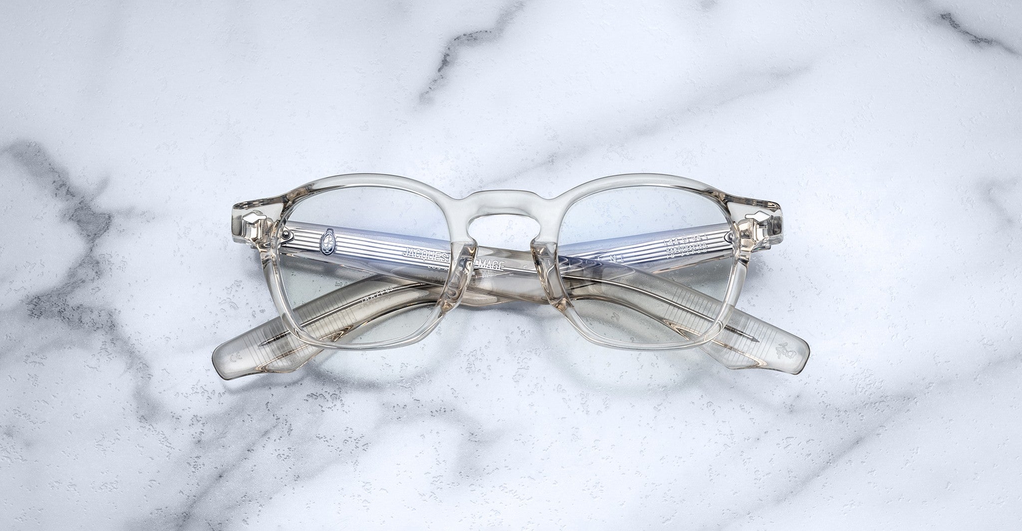 The Jacques Marie Mage Zephirin eyeglasses by Jacques Marie Mage, featuring round lenses and translucent frames, are folded and set on a white marble surface.