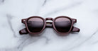 A pair of Jacques Marie Mage Zephirin round sunglasses in dark brown with tinted lenses rests on white marble, radiating timeless luxury and reflecting the exclusive style of limited edition Jacques Marie Mage eyewear.