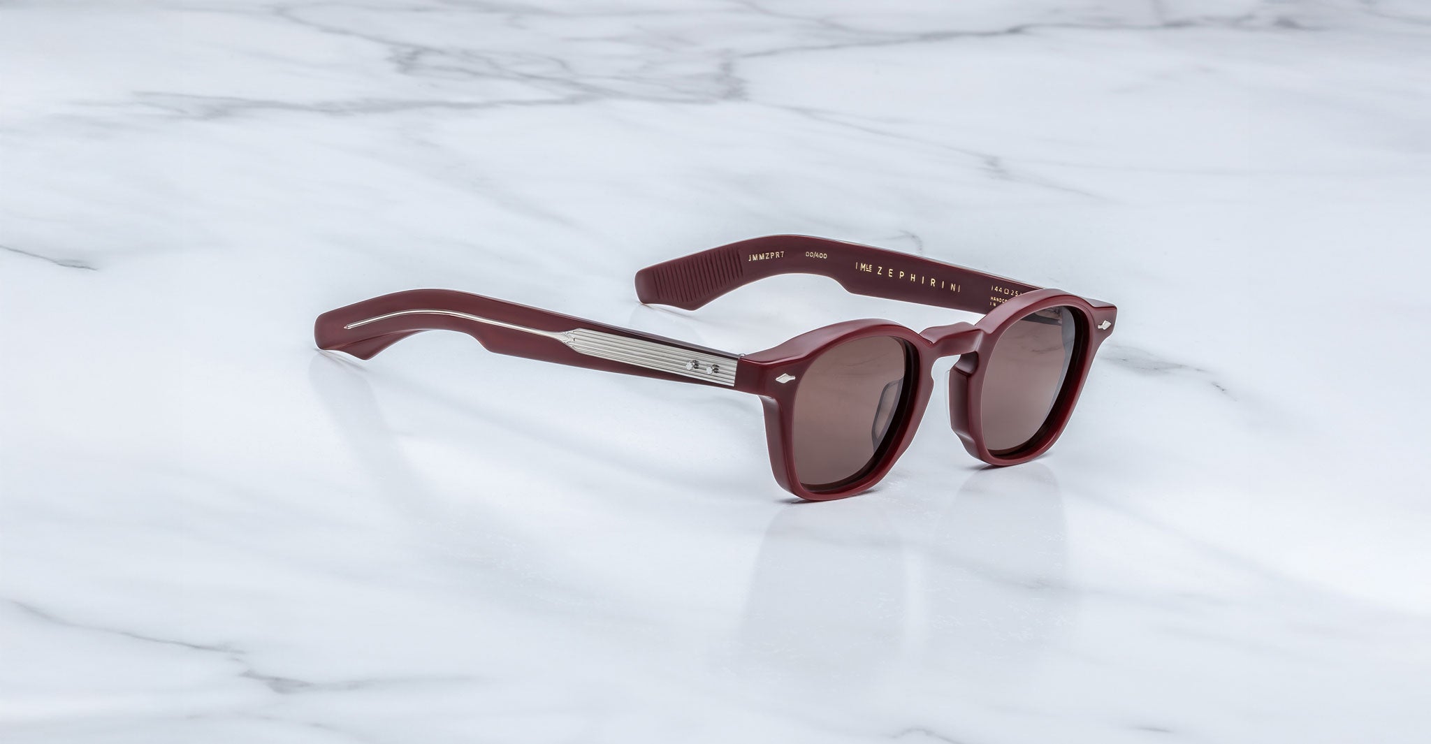 The Jacques Marie Mage Zephirin sunglasses by Jacques Marie Mage feature round maroon lenses, metallic arm accents, and a limited edition-inspired design, elegantly displayed on a white marble surface.