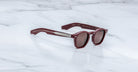 The Jacques Marie Mage Zephirin sunglasses by Jacques Marie Mage feature round maroon lenses, metallic arm accents, and a limited edition-inspired design, elegantly displayed on a white marble surface.