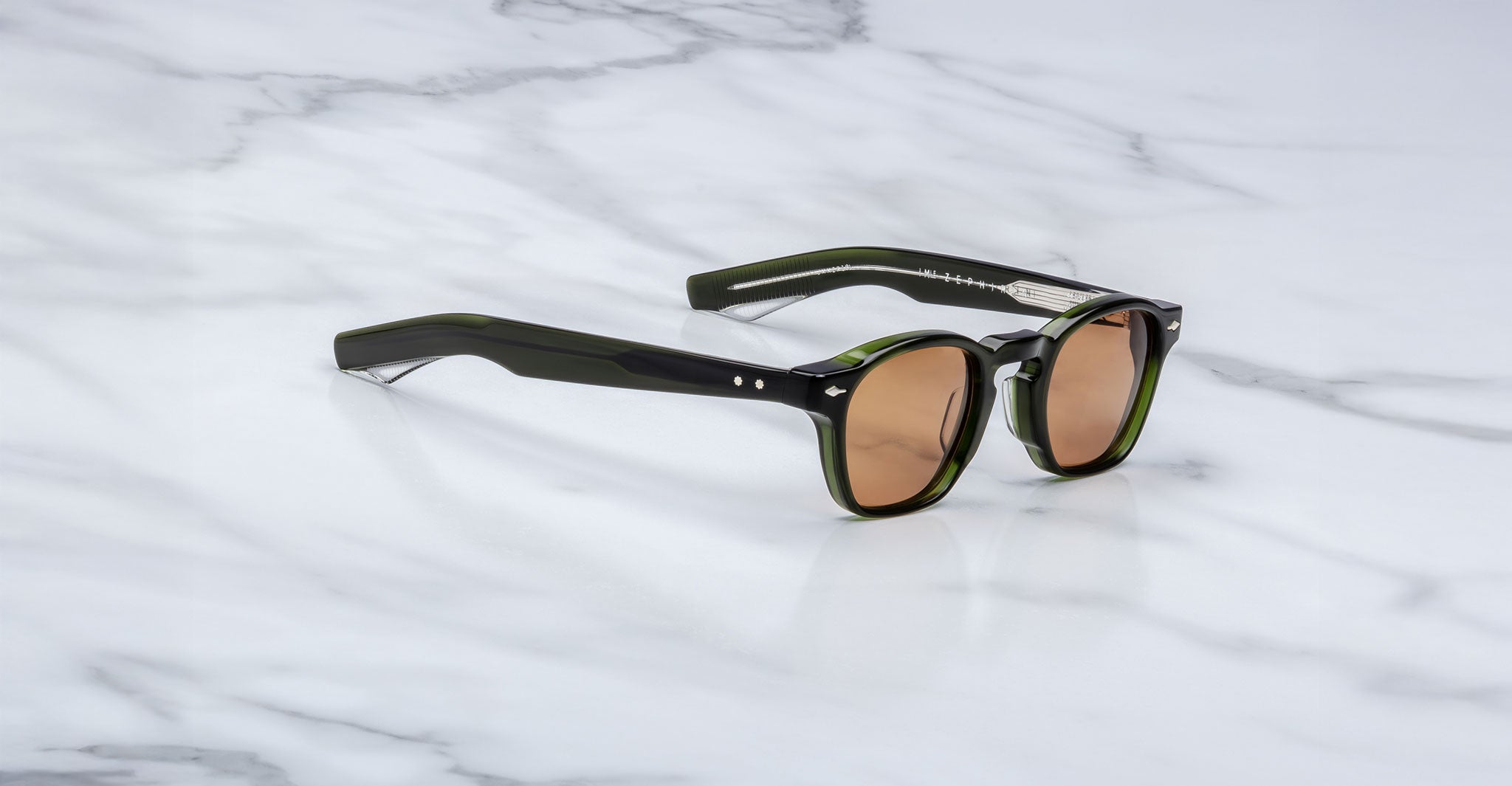 A pair of limited edition Jacques Marie Mage Zephirin sunglasses in dark green with brown lenses, crafted from premium acetate, rests on a white marble surface.