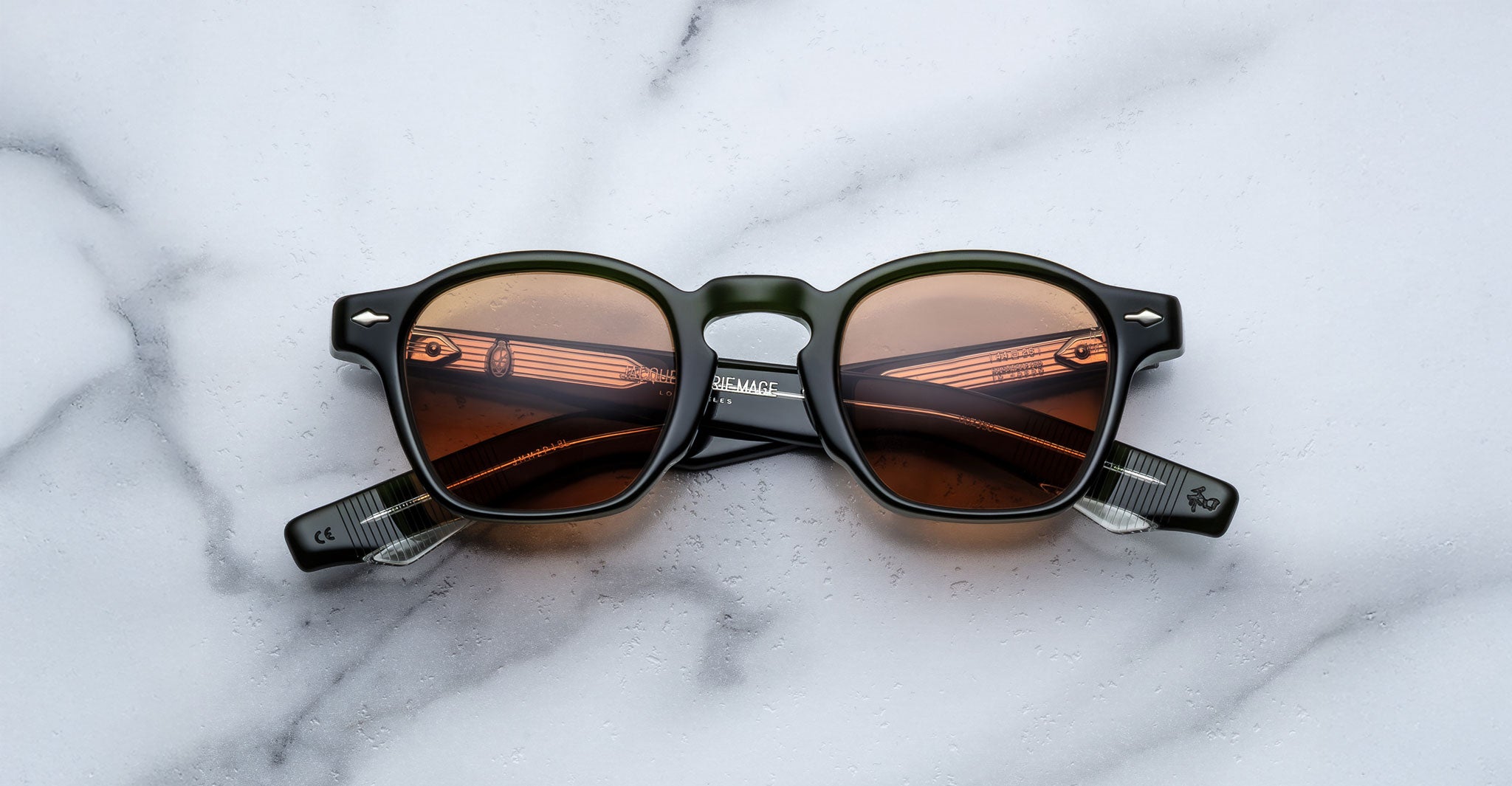 A pair of Jacques Marie Mage Zephirin sunglasses with round dark frames and brown-tinted lenses rests on white marble, highlighting the charm of this limited edition Jacques Marie Mage eyewear.