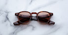 The Jacques Marie Mage Zephirin sunglasses by Jacques Marie Mage feature a brown tortoiseshell frame with red-tinted lenses, inspired by handcrafted acetate designs, displayed on a white marble surface.