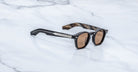Jacques Marie Mage Zephirin sunglasses in Brun, three-quarter view with dark brown acetate, warm brown lenses, and silver-tone accents on a white marble surface.