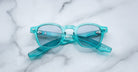 A pair of Jacques Marie Mage Zephirin turquoise translucent handcrafted acetate spectacles with clear lenses rests closed on a white marble surface.