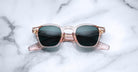 The Jacques Marie Mage Zephirin by Jacques Marie Mage features transparent pink handcrafted acetate frames with dark lenses, displayed on a white marble surface.