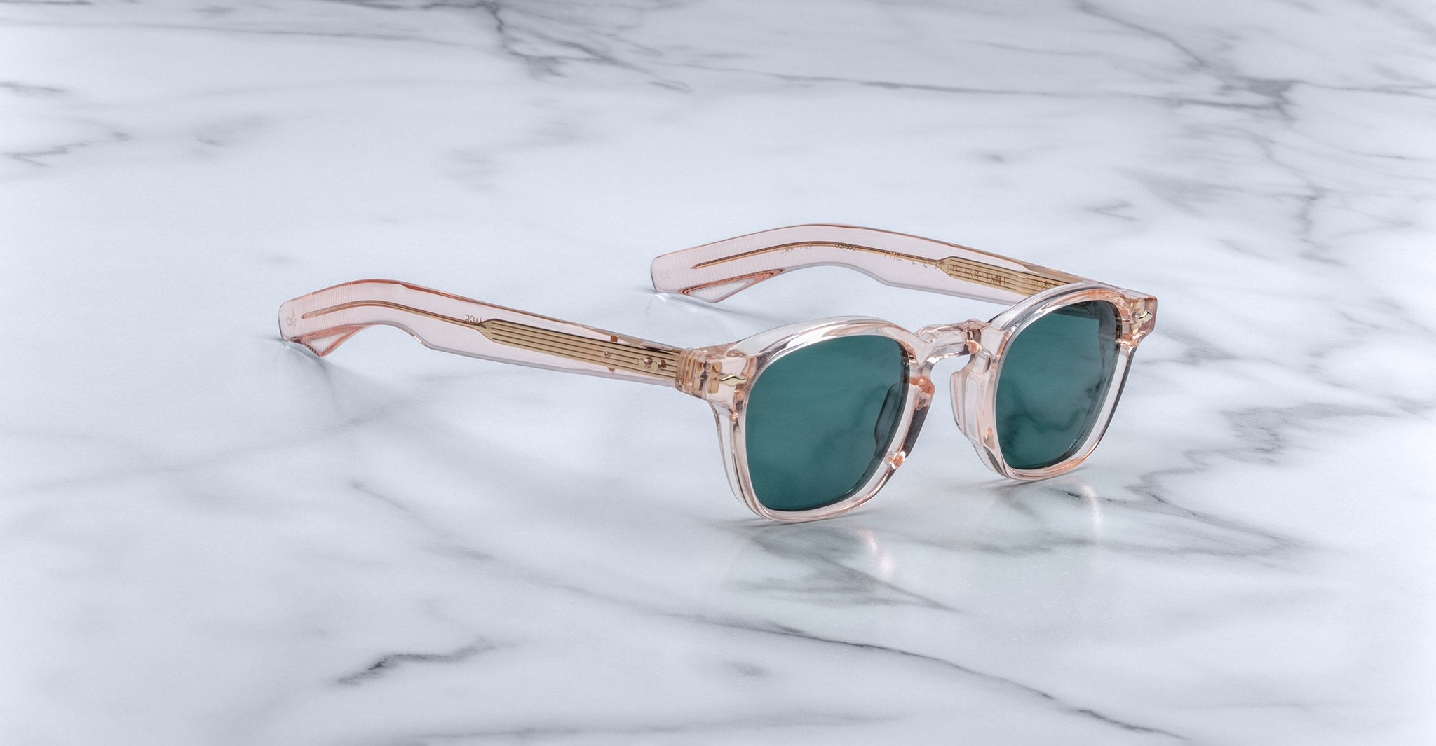 A pair of handcrafted Jacques Marie Mage Zephirin sunglasses in translucent pink with green lenses rest on a white marble surface, showcasing their limited edition appeal.